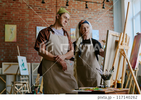 Medium long shot of senior artist helping young man to correct mistake during painting class in studio 117140262