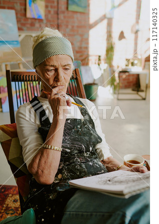 Vertical medium shot of pensive senior female artist sitting on chair with pencil in hand looking at sketch in sketchbook 117140325