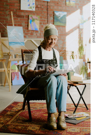 Vertical long shot of elderly Caucasian woman wearing apron sitting on chair in art studio and drawing in sketchbook 117140328