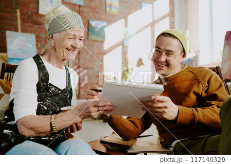 Creative young man showing sketch to cheerful senior art teacher during illustration class in studio 117140329