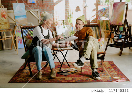 Senior Caucasian female artist sitting on chair in studio showing drawing in sketchbook to young man Senior Caucasian female artist sitting on chair in studio showing drawing in sketchbook to young man 117140331
