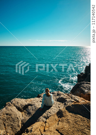 A girl sits on the edge of a cliff and looks at the sea A girl sits on the edge of a cliff and looks at the sea 117140542