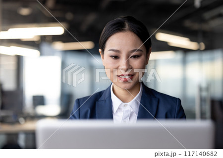 Smiling businesswoman working on laptop in modern office environment. Professional woman in formal attire focused on computer screen, representing productivity and corporate culture in workplace. 117140682