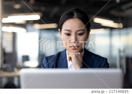 Businesswoman concentrating on work, using laptop in modern office. Professional attire, thoughtful expression. Corporate environment, productivity, focus. Businesswoman concentrating on work, using laptop in modern office. Professional attire, thoughtful expression. Corporate environment, productivity, focus. 117140685