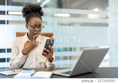 Businesswoman looking shocked while checking smartphone in modern office. Surprised expression on woman's face while sitting at desk with open laptop and documents. Businesswoman looking shocked while checking smartphone in modern office. Surprised expression on woman's face while sitting at desk with open laptop and documents. 117141055