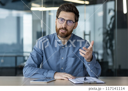 Distance Learning. Young serious man sitting at the desk in the office and conducting an online lecture, business training and meeting. Look and talk to the camera. 117141114