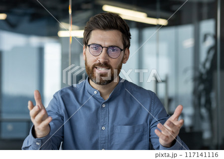 Close-up portrait of a young man in glasses and a blue shirt sitting in the office in front of the camera and talking on a video call, gesturing with his hands. 117141116