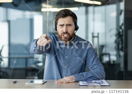 Portrait of a young man in a headset sitting in the office at the table, talking emotionally online, shouting and pointing at the camera. 117141156