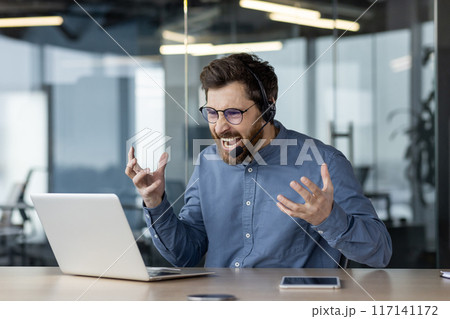 Aggressive young man wearing a headset sits in the office at a desk and talks unhappily on a video call, gesturing emotionally with his hands. 117141172