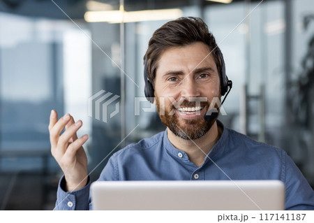 Happy businessman wearing headset smiling during video call in office. Confident man with laptop engaging in virtual meeting. Remote work, customer support and communication concept. 117141187