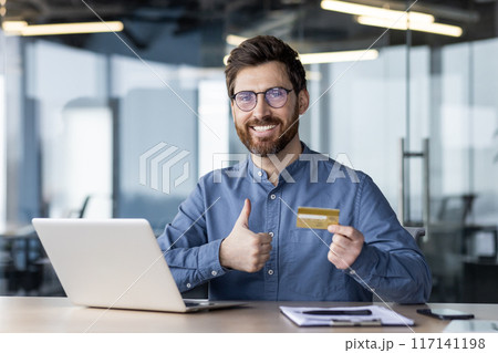 Portrait of a young smiling man sitting in the office at the table, holding and showing a golden credit card in his hand, showing the camera a super success sign with his finger. Portrait of a young smiling man sitting in the office at the table, holding and showing a golden credit card in his hand, showing the camera a super success sign with his finger. 117141198