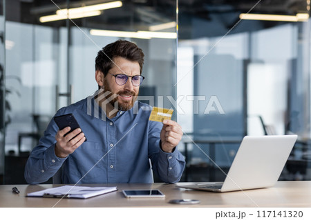 Worried young man sitting in the office at the desk, holding the phone in his hand and looking disappointedly at the plastic bank credit card. Problems with the account. Worried young man sitting in the office at the desk, holding the phone in his hand and looking disappointedly at the plastic bank credit card. Problems with the account. 117141320