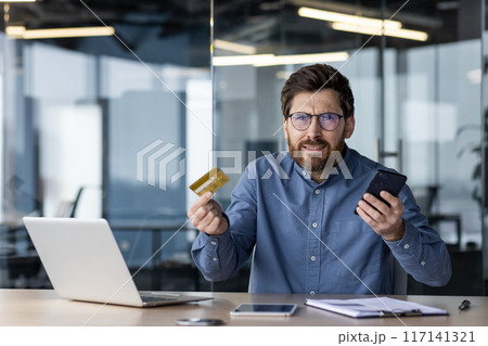 Frustrated man sitting in modern office holding credit card and smartphone, possibly dealing with online banking issues or fraud. Expression of confusion and concern while working with laptop 117141321