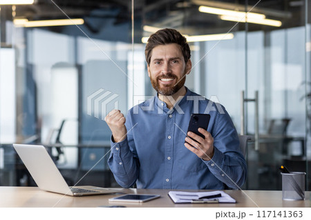 Portrait of a happy young businessman sitting at a desk in the office and looking at the camera, holding a mobile phone and showing a victory and success gesture with his hand. 117141363