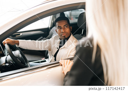 Back view of unrecognizable female auto dealer having communication with black male customer sitting in car salon. Professional saleswoman helping African-American client to choose best automobile. Back view of unrecognizable female auto dealer having communication with black male customer sitting in car salon. Professional saleswoman helping African-American client to choose best automobile. 117142417