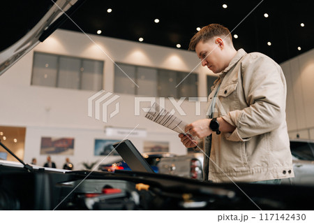 Shooting from below of thoughtful male customer holding papers, checking vehicle characteristics during choosing car in modern dealership center. Concept of choosing and buying new auto at showroom. Shooting from below of thoughtful male customer holding papers, checking vehicle characteristics during choosing car in modern dealership center. Concept of choosing and buying new auto at showroom. 117142430