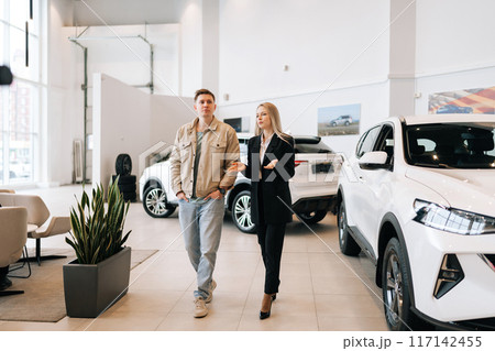 Wide shot of stylish female manager in suit showing new cars to male buyer and walking in modern showroom spbas. Young woman car dealer talking to man client and showing models of transport. Wide shot of stylish female manager in suit showing new cars to male buyer and walking in modern showroom spbas. Young woman car dealer talking to man client and showing models of transport. 117142455