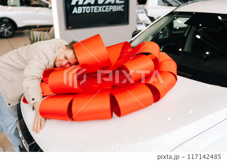 Joyful male customer lying on white luxury new car with large red bow on hood and smiling with closed eyes. Expensive purchase of cheerful male client person. Concept of buying new auto at showroom. 117142485