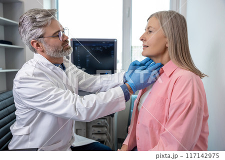 Male doctor palping female patient throat and looking focused Male doctor palping female patient throat and looking focused 117142975
