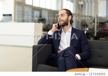 Salesperson in suit having phone conversation while sitting on sofa in contemporary car showroom Salesperson in suit having phone conversation while sitting on sofa in contemporary car showroom 117143030