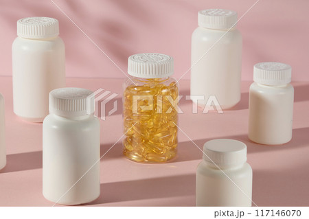 Neatly arranged medicine bottles mockup on pink counter top, transparent supplement bottle filled with yellow vitamin capsule surrounded in center by white unbranded bottles. High angle, copy space 117146070