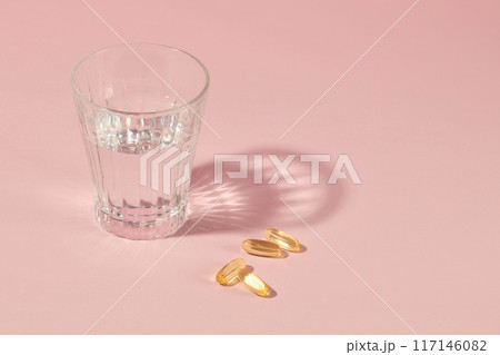 Nutrient supplement products advertising with minimalist style with a glass of water and a few flat lay yellow capsules, featured on plain pink surface. Displaying space, high angle view 117146082