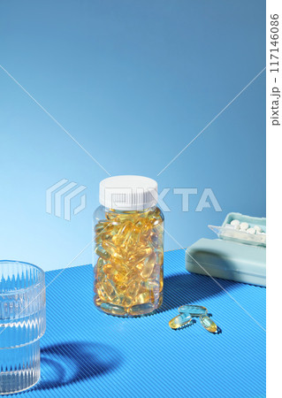 Blue background photo was taken from high angle view. A clear jar filled with yellow supplement capsules, a daily pill box and a glass of water prepared for medicine drinking Blue background photo was taken from high angle view. A clear jar filled with yellow supplement capsules, a daily pill box and a glass of water prepared for medicine drinking 117146086
