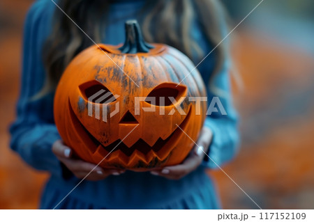 Woman in a blue dress holding a sinister carved pumpkin, capturing a moody Halloween vibe 117152109
