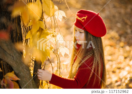 Cute girl in a burgundy beret stands near a tree in autumn b touches a yellow leaf Cute girl in a burgundy beret stands near a tree in autumn b touches a yellow leaf 117152962