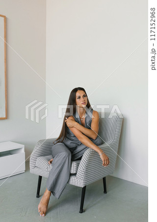Young woman is sitting in a chair wearing a grey outfit. She is wearing a necklace and has her arms crossed 117152968