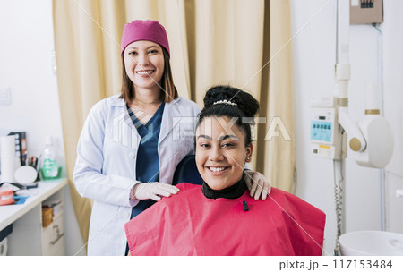 Female dentist with satisfied patient smiling at camera, Female dentist with patient smiling friendly. Portrait of dentist with patient smiling at camera in office 117153484