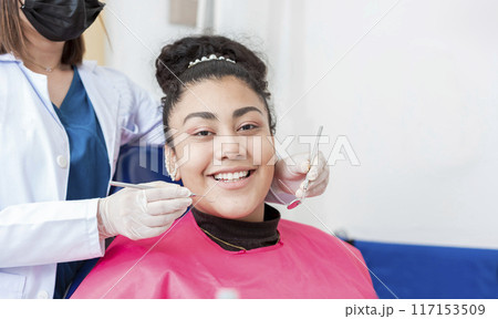 Dentist doctor examining mouth to female patient looking at camera lying on chair, Female dentist with probe and dental mirror examining mouth to lying patient. Health and dental care concept 117153509