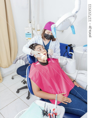 Dentist in his office examining female patient's mouth. Female dentist examining mouth to teen girl lying on chair, Female dentist with probe and dental mirror examining young female mouth 117153511