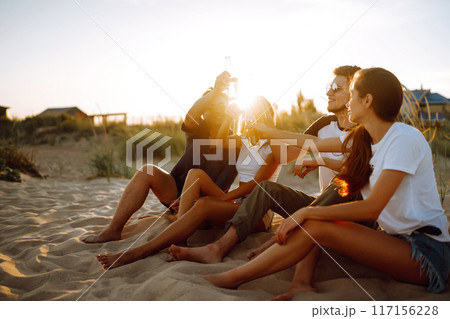 Young people sitting together at beach, drinking beer. Group of friends cheers with beers at the beach. 117156228