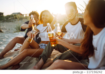 Young people sitting together at beach, drinking beer. Group of friends cheers with beers at the beach. Young people sitting together at beach, drinking beer. Group of friends cheers with beers at the beach. 117156242