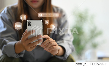 Young woman using smartphone, interacting with various multimedia icons including music, video and gaming Young woman using smartphone, interacting with various multimedia icons including music, video and gaming 117157027