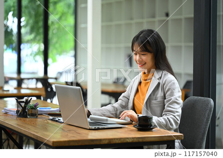 Asian businesswoman sitting at desk with laptop, using a calculator and analyzing financial documents Asian businesswoman sitting at desk with laptop, using a calculator and analyzing financial documents 117157040