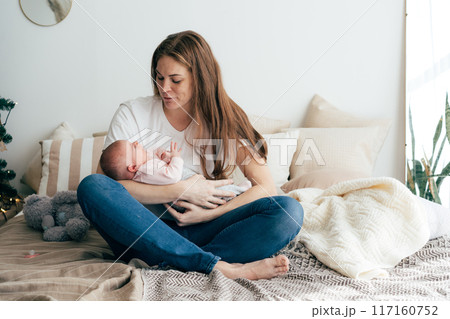 A tender caring mother nurses a newborn baby in her arms while sitting on the bed. A tender caring mother nurses a newborn baby in her arms while sitting on the bed. 117160752