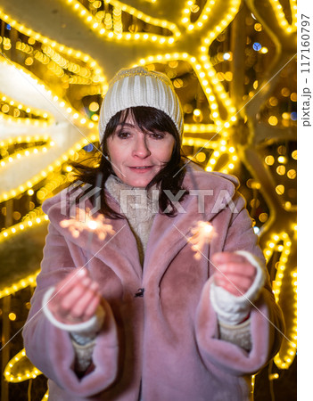 Russian 48 years old woman in blue sweater and pink coat standing against yellow christmas lights before Christmas and holding lit sparklers 117160797