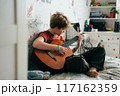 Romantic adolescent redhead girl learning to play the guitar while sitting in the room.	 117162359