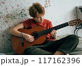 Romantic adolescent redhead girl learning to play the guitar while sitting in the room.	 117162396