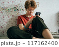 A teenage girl sitting on a bed in a room uses a mobile phone for online communication.	 117162400