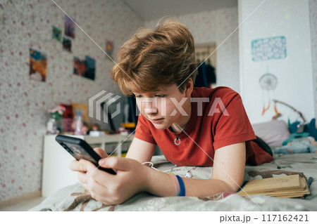 A teenage girl lying on a bed in a room uses a mobile phone for online communication. A teenage girl lying on a bed in a room uses a mobile phone for online communication. 117162421