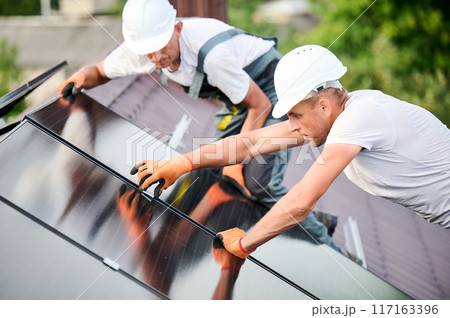 Workers building solar panel system on rooftop of house. Two men installers in helmets installing photovoltaic solar module outdoors. Alternative, green and renewable energy generation concept. Workers building solar panel system on rooftop of house. Two men installers in helmets installing photovoltaic solar module outdoors. Alternative, green and renewable energy generation concept. 117163396
