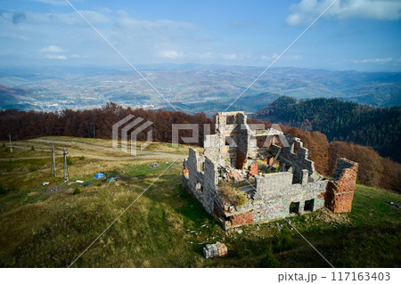 Decaying stone building on grassy hill, overlooking landscape of mountains and valleys. Ruins contrast with vibrant autumnal forest and distant village below, under bright blue sky with clouds. 117163403