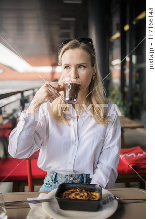 Blonde gen z woman eating lasagna and drink cocoa in the cafe on break. Gen z girl spend time in restaurant 117166148
