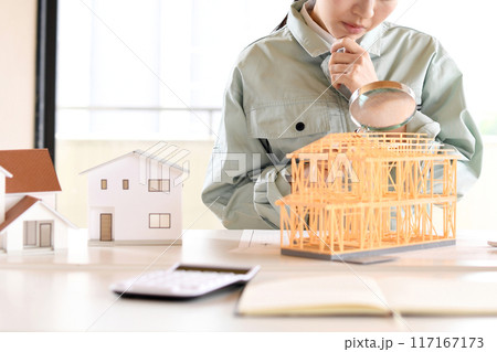 住宅模型を虫眼鏡で覗く作業服姿の若い女性 117167173