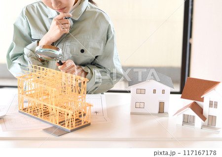住宅模型を虫眼鏡で覗く作業服姿の若い女性 117167178