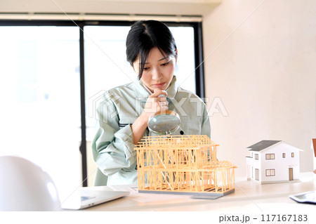 住宅模型を虫眼鏡で覗く作業服姿の若い女性 117167183