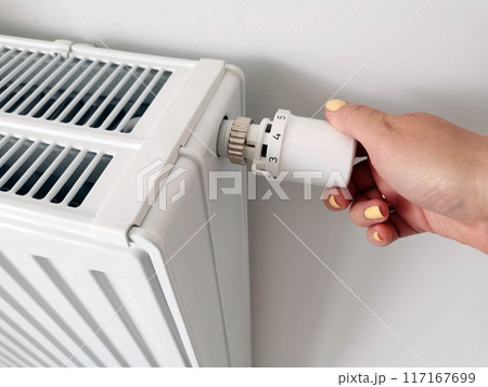 A woman's hand regulates the temperature in the room with a white radiator thermostat on a white wall background. Control of temperature and climatic conditions in an apartment, house, office A woman's hand regulates the temperature in the room with a white radiator thermostat on a white wall background. Control of temperature and climatic conditions in an apartment, house, office 117167699